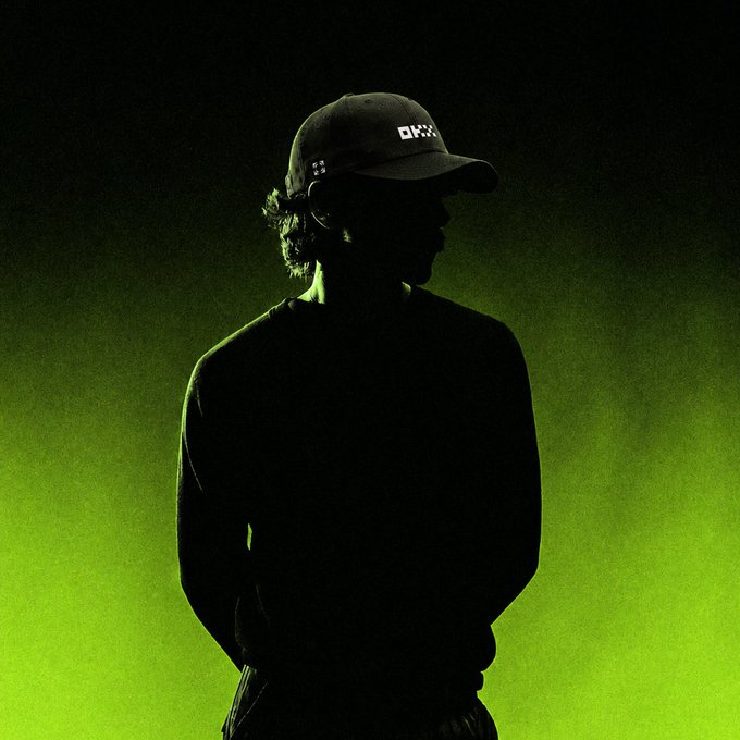 A moody silhouette of a person wearing a dark hoodie and a cap with 'OXXX' written on it, standing against a glowing green smoky background.