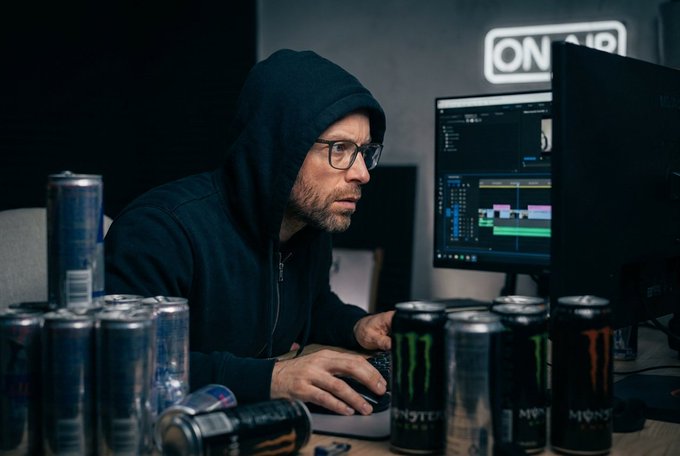 A man in a black hoodie and glasses working at a computer with video editing software open, surrounded by numerous empty Red Bull and Monster energy drink cans in a dimly lit room with an ON AIR sign in the background.