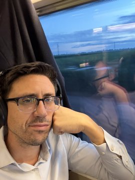 A man wearing glasses resting his chin on his hand while looking out a train window at dusk, with his reflection visible in the glass.