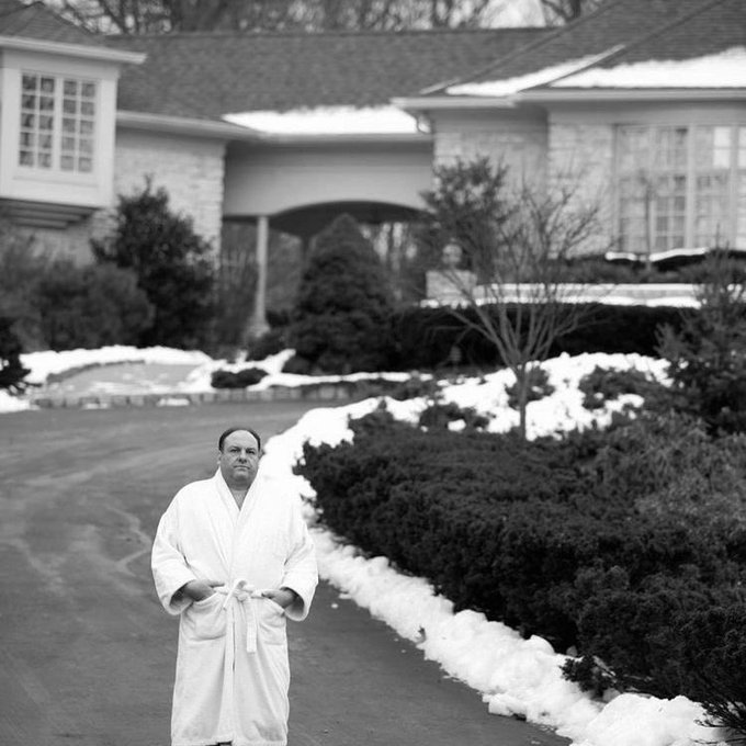 A black and white photo shows a balding man in a white bathrobe standing on a snowy driveway in front of a large house.