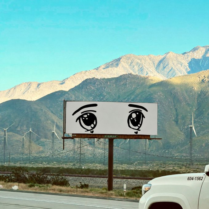A billboard displaying large anime-style eyes with tears, set against a backdrop of mountains and wind turbines, with a white truck marked '604-1562' partially visible in the foreground.
