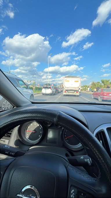 A driver's perspective from inside a car showing the steering wheel and dashboard, with a view of traffic on a highway under a bright blue sky with scattered clouds.