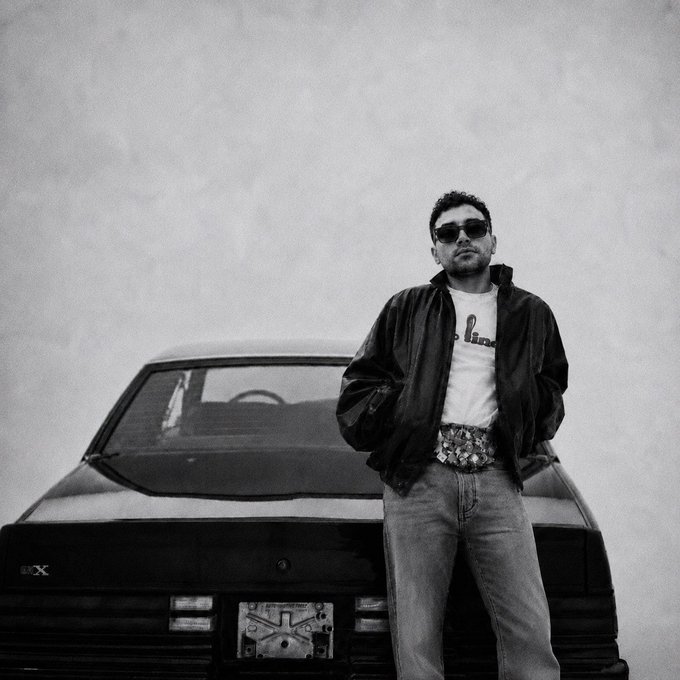A black and white photo of a man wearing sunglasses, a t-shirt, jacket, and large belt buckle standing in front of a sports car with a 'DX' badge visible on the rear.