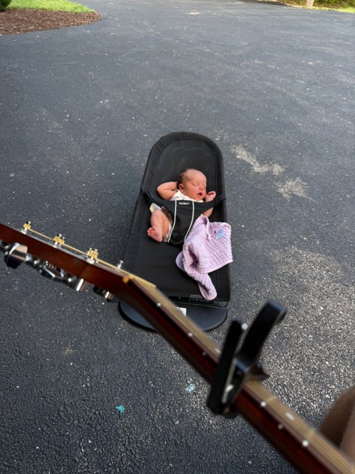A baby lying in a black baby lounger on an asphalt surface, with a guitar neck visible in the foreground, suggesting someone is playing music for the baby.