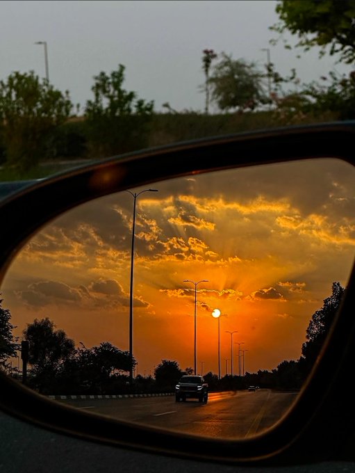An artistic photograph of a sunset sky with orange clouds reflected in a car's side mirror, with street lights and a vehicle on the road visible in the reflection.