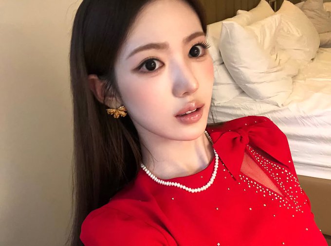 The image shows a glamorous selfie of a young woman with long dark hair, wearing a striking red dress adorned with pearls and a large bow, along with a delicate pearl necklace and gold floral earrings. The hashtags #SUI, #KiiiKiii, #수이, and #키키 suggest this is KiiiKiii member Sui (Choi Sui), a South Korean girl group, posing elegantly in a hotel or bedroom setting with white bedding visible behind her.