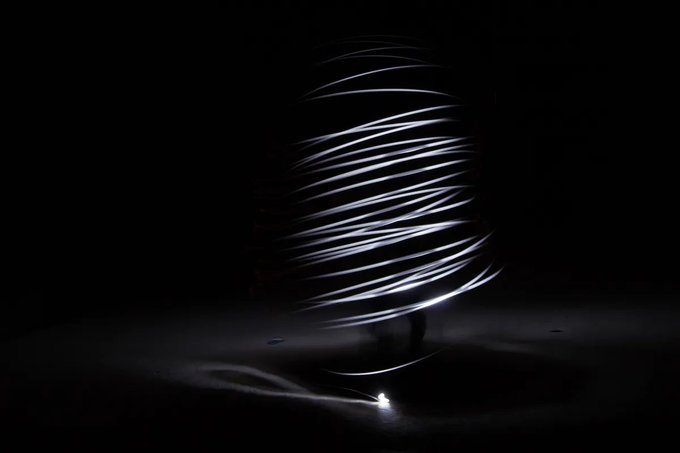 Light trails spiral around a figure in a dark setting, creating an abstract long-exposure effect with a small light source at the base.