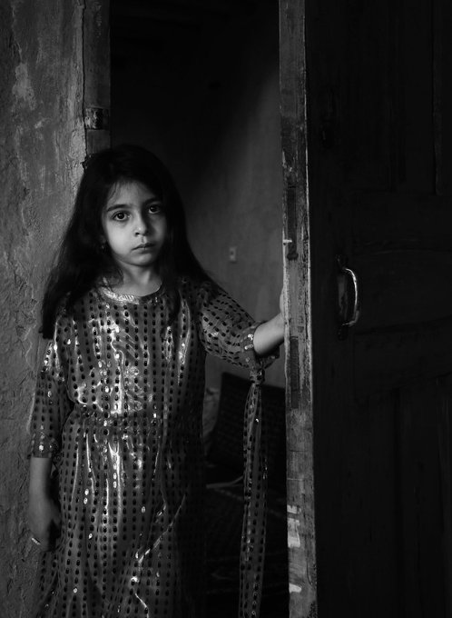A young girl in a sequined dress stands in a dimly lit doorway, gazing out with a solemn expression—capturing the haunting, nostalgic mood of "Grandma's house."