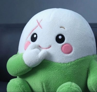 A plush toy with a white head and green body has a pink 'X' marked on its forehead, blushing cheeks, and its hands clasped near its mouth in a shy or giggling pose.
