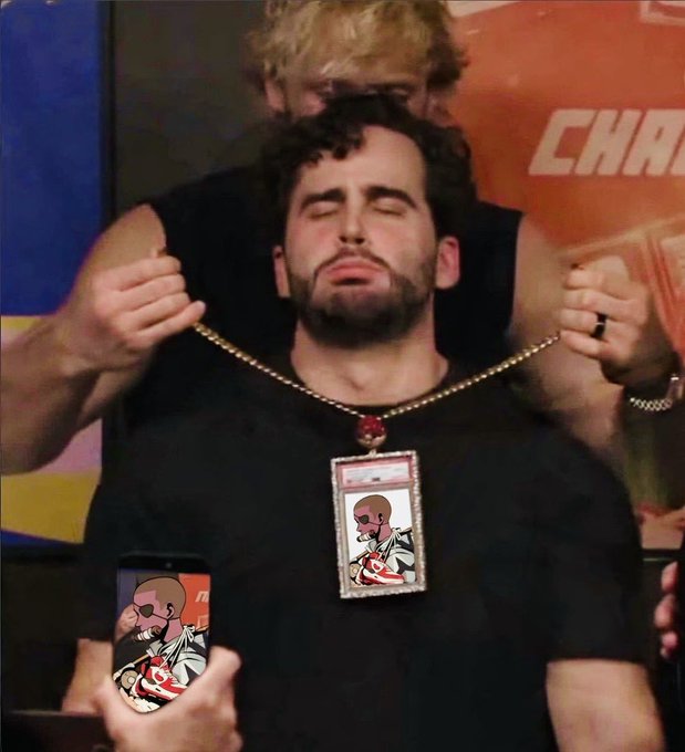 A man with dark curly hair and a beard wearing a black shirt has his eyes closed as someone places a large jeweled chain with a pendant around his neck, with another person filming the moment on a phone showing a cartoon avatar.