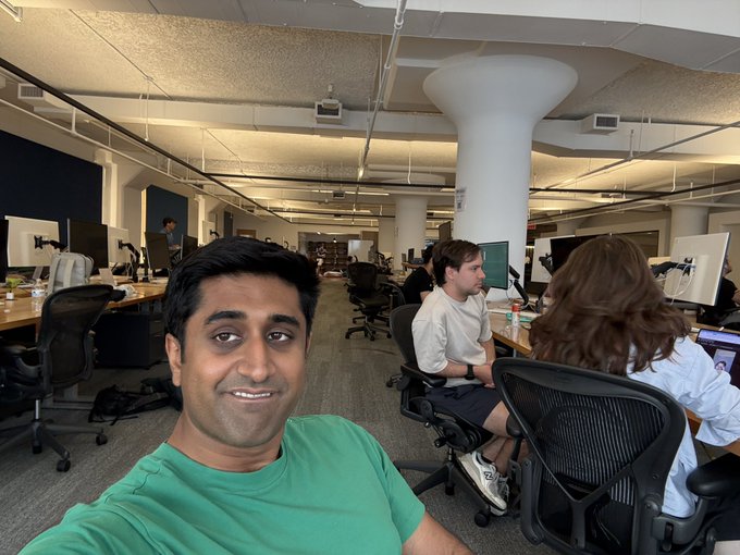 A man in a green t-shirt takes a selfie in a bustling modern office space filled with coworkers at their desks and computer monitors, capturing the lively "blockchain energy" he mentions while working from Monad. The open-floor workspace with its high ceilings and rows of workstations visually reflects the active, collaborative environment described in the post.