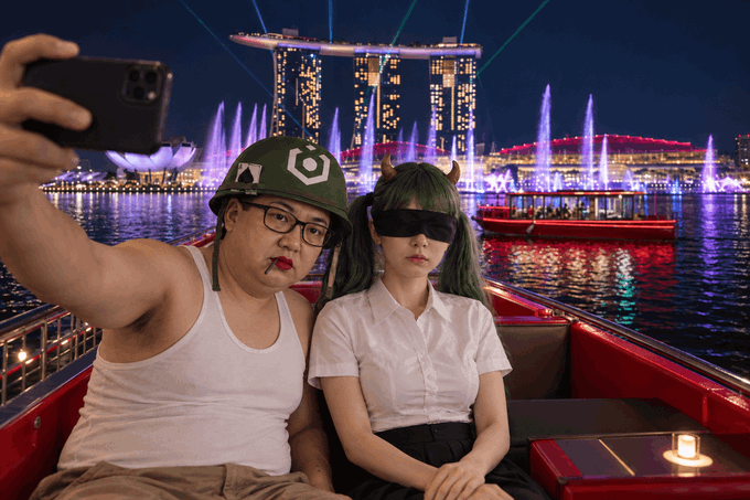 A couple taking a selfie while seated on a boat at night; the man wears a military-style helmet and glasses, while the woman has dark hair with cat ears, with a colorful fountain show and city skyline (resembling Marina Bay Sands in Singapore) visible in the background.