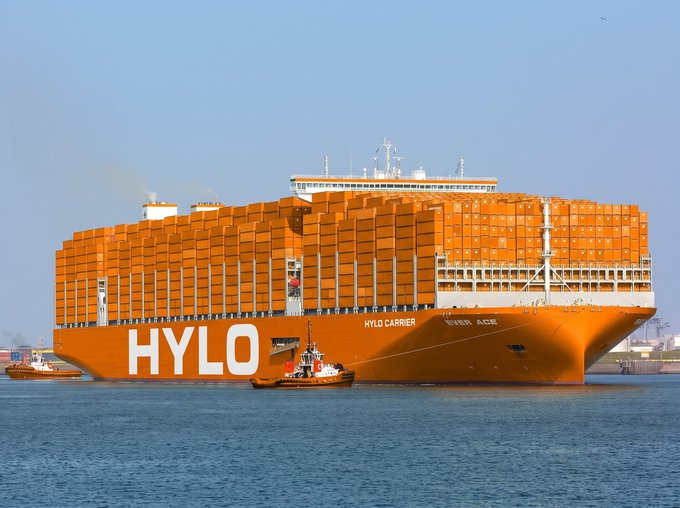 A large orange HYLO container ship named 'EVER ACE' being guided by a small tugboat in a harbor under clear blue skies.