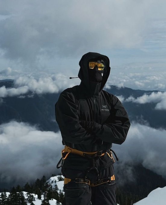 A person dressed in a black hooded Arc'teryx jacket and yellow-tinted goggles stands with crossed arms on a snowy mountain peak, with clouds and mountain ridges behind them.