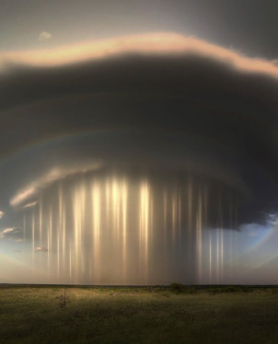 A dramatic landscape showing a massive dark cloud formation with streaks of rain or light falling from it over a flat grassy plain.