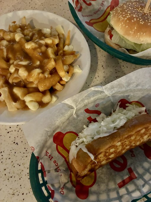 An overhead photo of a fast-food meal featuring poutine (fries with gravy and cheese curds) on a white plate, a burger on a tray, and what appears to be a Vietnamese-style sandwich on branded wrapper paper.