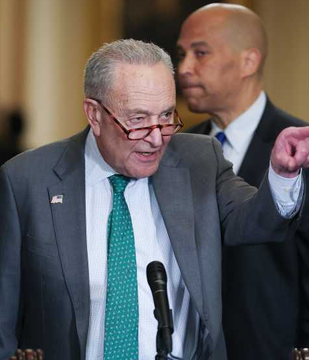 Senate Minority Leader Chuck Schumer, wearing glasses and a green patterned tie, speaks passionately into a microphone while gesturing with his hand, as he introduces an anti-corruption bill targeting alleged Trump-related crypto profits. Another man in a suit stands behind him in what appears to be a Capitol Hill setting.