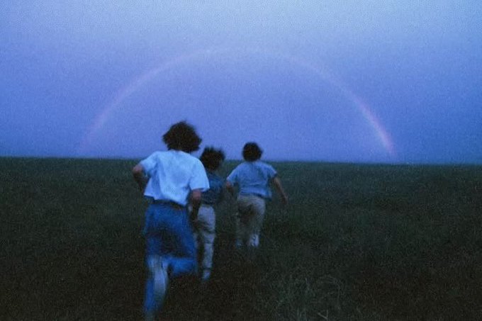 Three people run across a vast grassy field toward a large rainbow arching across the twilight sky, capturing the hopeful, forward-moving spirit of the "Good morning everyone, let's have a productive day" message as they chase the dawn's promise.