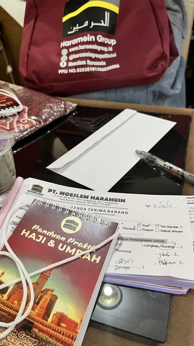 Photo showing a travel agency setup with a 'PT. Mabrur Haromain' bag, a 'Panduan Praktis Haji & Umrah' booklet, order forms dated 16/12/16, and a pen.