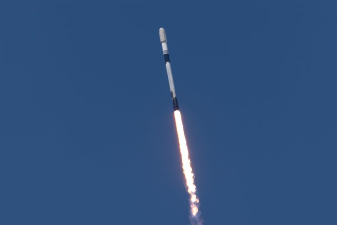 A SpaceX rocket ascends through a clear blue sky, leaving a bright trail of exhaust behind it.