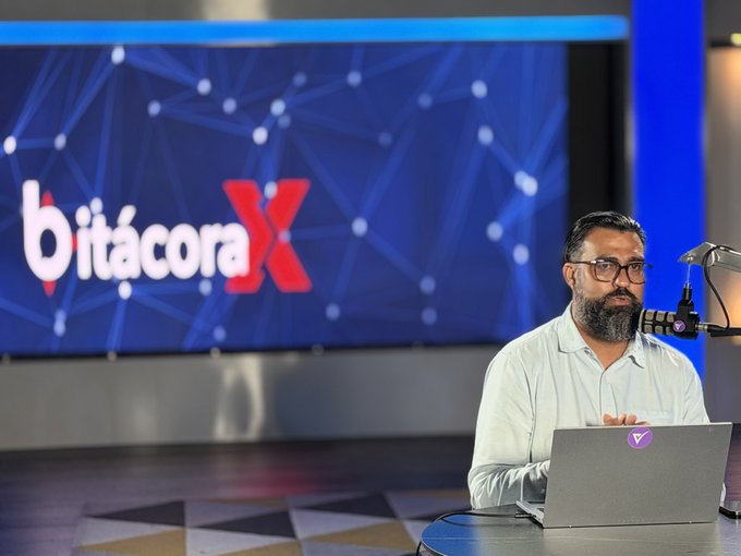 A man with a beard and glasses, wearing a light-colored shirt, sits at a desk with a laptop and microphone in front of a large screen displaying 'Bitácora X'.