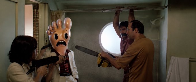 A scene from a film showing a person wearing a foot-shaped mask being threatened with a saw and a weapon in a dimly lit room with a round window.