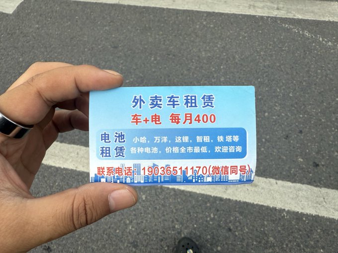 A hand holds a blue business card advertising food delivery vehicle rentals for 400 yuan per month (car and battery), with battery rentals from various brands, listing contact number 19036511170. The card, picked up off the street, humorously serves as evidence of the poster's "liquidation" after losing their food delivery job, as they now face handing out these rental flyers for new gig work.
