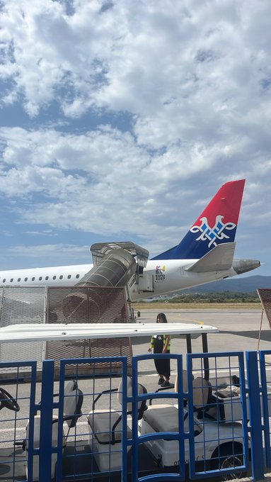 A photograph taken at an airport shows the tail of an airplane with a red and blue livery featuring a lion logo, with a mobile staircase attached and a worker in a yellow vest nearby.