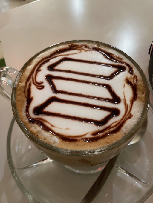 The image features a cup of hot chocolate with white swirls, likely made from whipped cream or caramel sauce, sitting inside a glass mug. The mug is placed on top of a saucer, which rests on a dining table. In the background, there are two blurred people who appear to be enjoying their time together at the table.