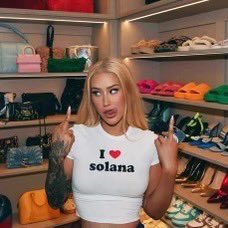 A woman standing in a walk-in closet filled with colorful handbags, wearing a white crop top that reads 'I ❤ solana'.