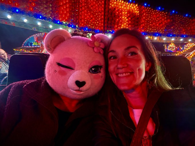 A selfie of a smiling woman posing next to a person wearing a pink teddy bear mascot head with a flower accessory, taken in front of colorful amusement park lights.
