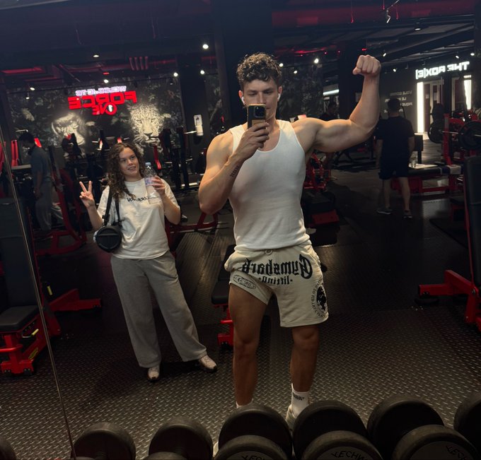 A muscular man in a white tank top and shorts takes a mirror selfie flexing his bicep in a gym, while a woman in the background poses with a peace sign.