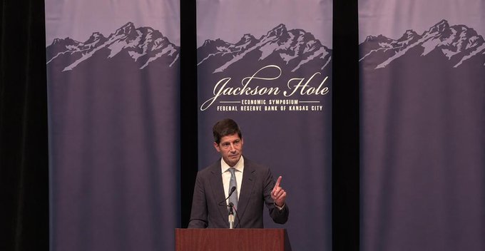 Fed Chair Kevin Warsh delivers a keynote address at the Federal Reserve Bank of Kansas City's Jackson Hole Economic Symposium, gesturing with his right index finger raised as he stands at a podium in front of banners displaying the event's mountain logo and "Jackson Hole Economic Symposium" text, discussing the Fed's commitment to its 2% PCE inflation target and ongoing price stability concerns.