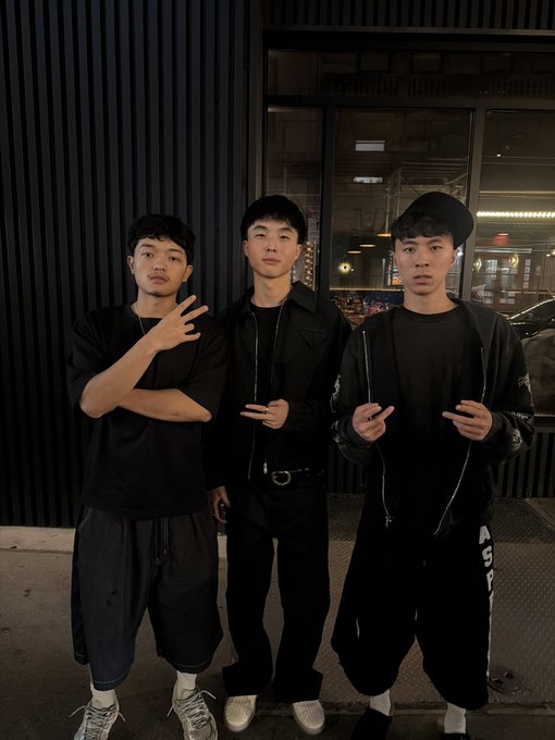 Three young men pose together at night in matching black streetwear outfits, with one wearing a cap and white earbuds, standing outside what appears to be a building entrance. The image aligns with the post's caption by visually portraying "Kimchi, Jasontheween, and Ray" as trendy, celebrity-like figures through their coordinated stylish appearance and confident poses.