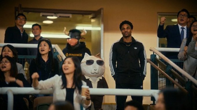 A group of people in a stadium-style setting cheering, with a person wearing a white dog mask and sunglasses standing among them.