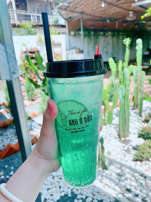 A hand holding a clear plastic cup filled with a green iced beverage from 'Anh Ố Ốu' coffee, set against an outdoor background with cacti.