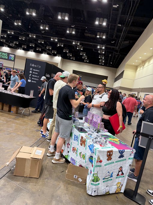 Attendees gather around a colorful VeeFriends-branded table at what appears to be a convention, engaging with merchandise and merchandise boxes while an attendee handles a product alongside staff members. The lively scene captures the growing community and brand presence Gary Vaynerchuk references, showcasing the energy and expansion of the VeeFriends movement from his earlier gatherings.