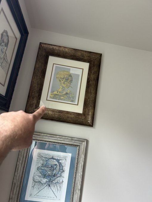 A first-person view of a hand pointing at framed artwork on a wall, with the central piece displaying a yellow skeletal portrait inside a textured bronze-colored frame.