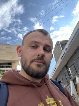 A selfie of a bearded man wearing a maroon hoodie, taken outdoors with a blue sky, power lines, and buildings in the background.