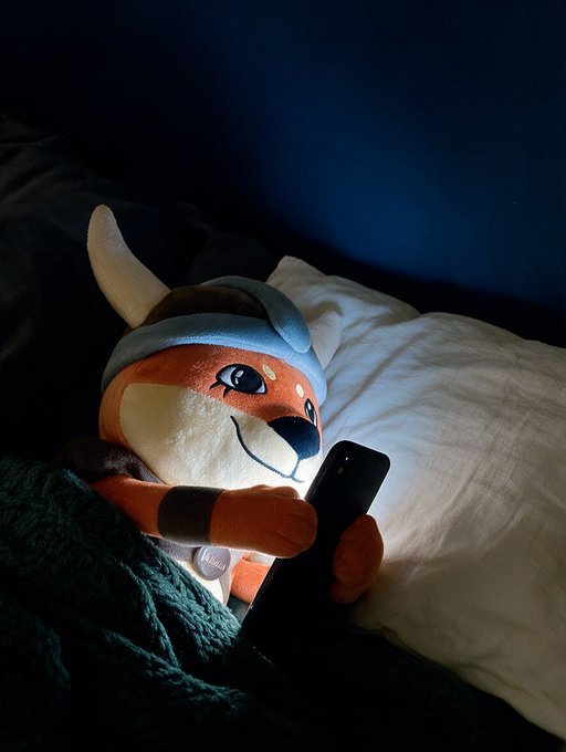 A plush toy of a character with a horn and helmet lying in bed under a blanket, holding a smartphone that illuminates its face.
