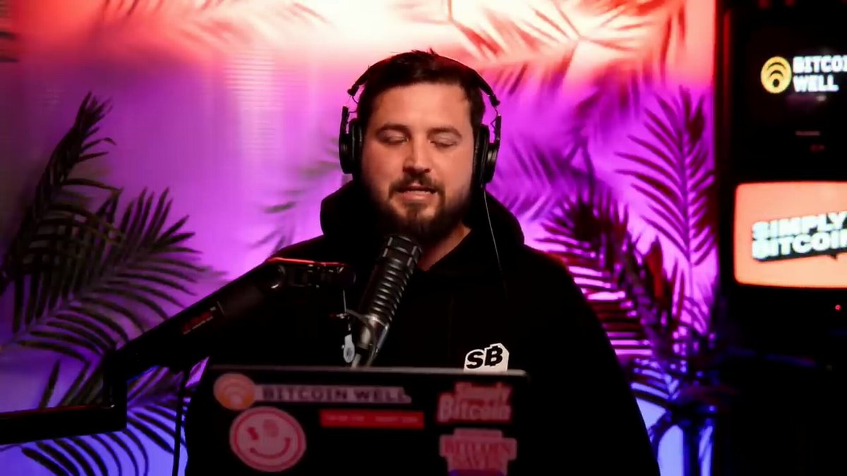 The image features a man wearing headphones while sitting at a desk with a laptop computer. The man appears to be speaking into a microphone, possibly participating in an online podcast or live stream. The background of the image includes a purple wall and a TV screen displaying "Bitcoin Well".