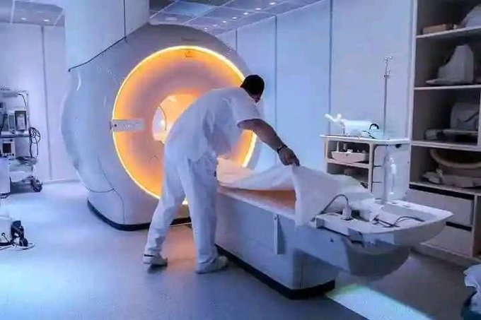 A medical worker in white clothing prepares a patient on a table in a clean clinical room with a large circular scanner glowing orange behind them.