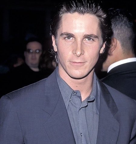 A vintage photograph of a young man with dark slicked-back hair wearing a gray suit jacket and button-up shirt, appearing to be at a formal event.