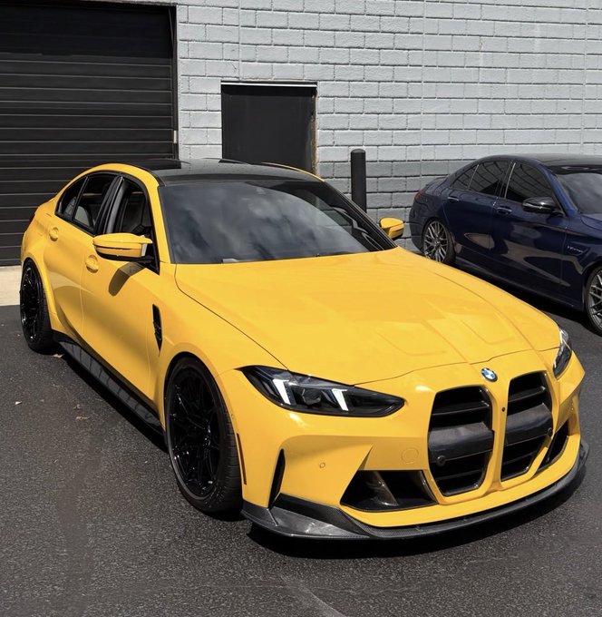 A bright yellow BMW M sedan is parked on asphalt in front of a gray brick building, with a dark blue car partially visible behind it.