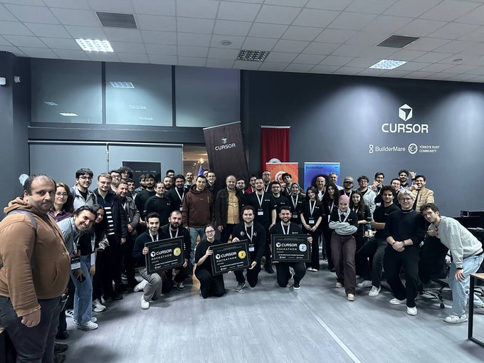 A large group of people poses for a group photo in front of a wall displaying the 'CURSOR' logo, with some individuals in the front row holding certificates labeled 'CURSOR MASTERMIND'.
