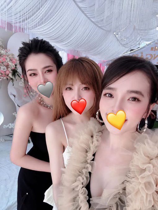 The image features three women posing together for a photo with their faces partially obscured by heart-shaped masks. The woman on the left wears a black dress, while the other two women are wearing ruffled dresses. They all have smiles on their faces as they pose for the picture.