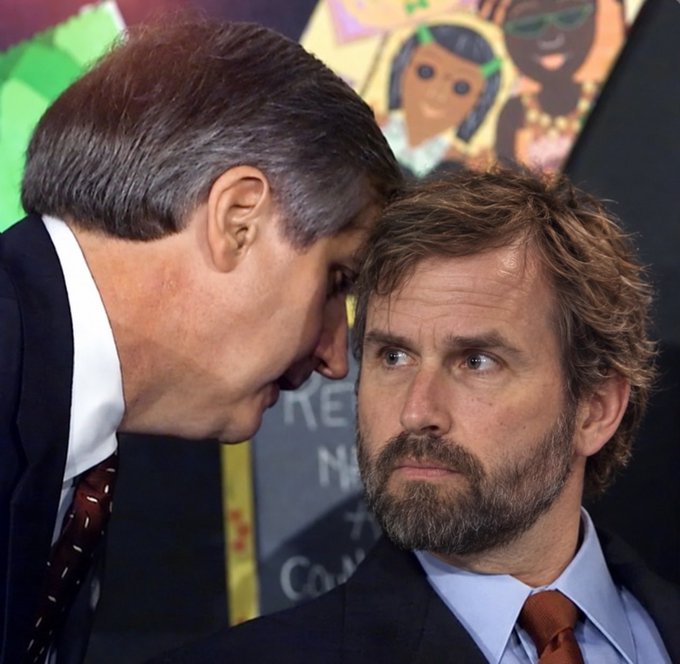 A man in a suit whispers into another man's ear while standing in front of a poster. The image appears to symbolize the hidden, confidential nature of a new privacy protocol being compared to Zcash and Bitcoin.