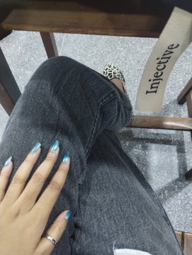 The image shows a person's hand resting on a chair, with their fingernails painted blue. The background of the photo features a wooden chair and a white sign that says "INJ" or "INJECTIVE". The caption at the bottom of the image reads "Touching grass = Going to Uni because its exam season but hey I'M always with my fav $INJ bag rate my nails btw".