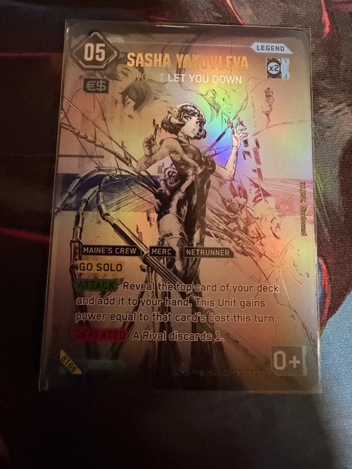 A holographic "Iconic Secret" foil trading card featuring Sasha Yakovleva titled "I Let You Down," numbered 05 and valued at $169, rests on a dark fabric surface—showcasing one of the rarest pulls from the Cyberpunk TCG Kickstarter boxes.