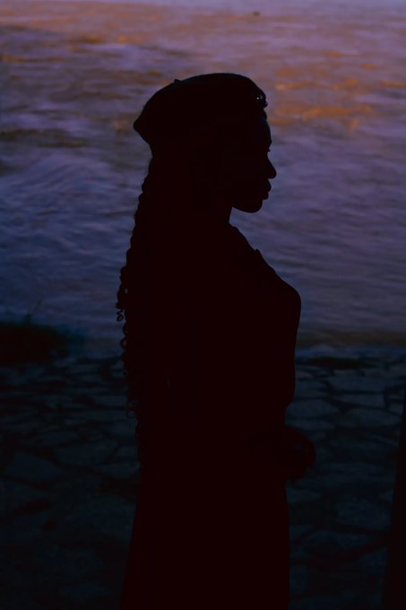 A silhouette of a person with long braided hair stands in profile against a softly lit body of water reflecting purple and orange hues at dusk.